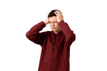 Handsome teenage boy suffering from headache or migraine, isolated on white background. Young guy wearing maroon hoodie wincing in pain holding his head in his hands feeling stressed and depressedの写真素材