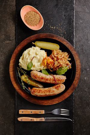 Traditional German Sausages with Mashed Potato and Sauerkraut. Wurst or Bratwurst with Fermented Cabbage, Pickled Cucumbers, and Spices in Rustic Wood Bowl on Natural Black Stone Background Top Viewの写真素材