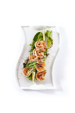 Hot Appetizers of Skewered Tiger Prawns Fried with Garlic on Elegant Restaurant Plate Isolated on White Background. Warm Starter with Grilled King Shrimps with Herbs, Spices, Lime and Greens Close Upの写真素材