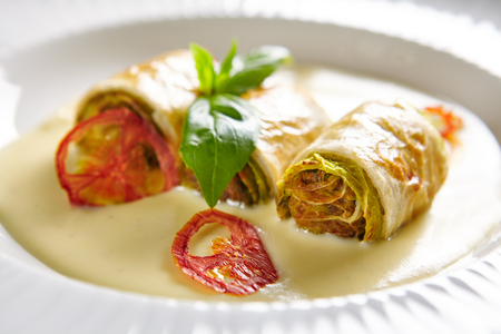 Macro Photo of Baked Mini Cabbage Rolls in Creamy Sauce on White Plate with Blurred Background. Exquisite Serving Vegetarian Dish with Stuffed Cole Leaves, Fresh Basil and Sun Dried Tomatoes Close Upの写真素材