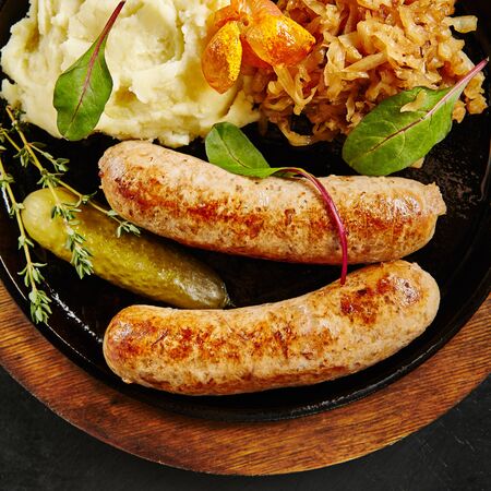 Traditional German Sausages with Mashed Potato and Sauerkraut. Wurst or Bratwurst with Fermented Cabbage, Pickled Cucumbers, and Spices in Rustic Wood Bowl on Natural Black Stone Background Top Viewの写真素材