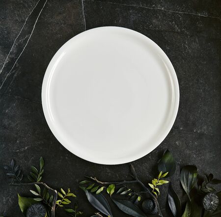 Exquisite serving white restaurant plate template on natural dark stone and leaves background top view with place for text. Blank porcelain dish mockup for your designの写真素材