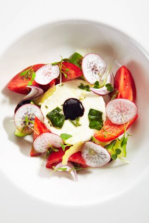 Exquisite serving salad of fresh vegetables, ricotta cheese and fragrant herbs on white restaurant plate isolated. High cuisine restaurant dish with delicious greek village salad top viewの写真素材