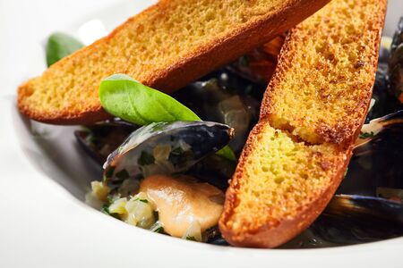 Mussel in white wine sauce closeup. Seafood with crunchy baguette slices in plate. Marine food with roasted bread and basil leaves meal ingredients. French dish isolated on white backgroundの写真素材