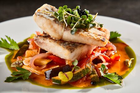 Pollock fish fillet with vegetables on white plate. Gourmet delicious food. Baked fish piece with sweet and sour sauce and herbs side view. Seafood with steamed veggies meal on black tableの写真素材