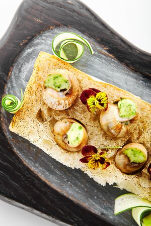Snails with garlic and cheese top view. Delicious escargots with sauce. Haute cuisine dish on wooden tray. Delicacy meal served with violets on bread. French food presentation, culinary menuの写真素材