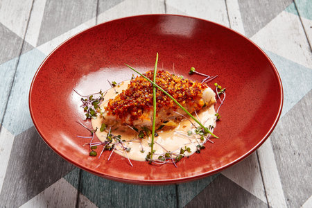 Cod piece with sauce in red deep plate. Fish cooked in tomato crumbs side view. Seafood dish in bowl...