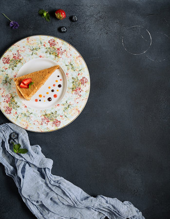 Sweet homemade layered honey cake on a black textured table with blue cloth and fresh strawberry