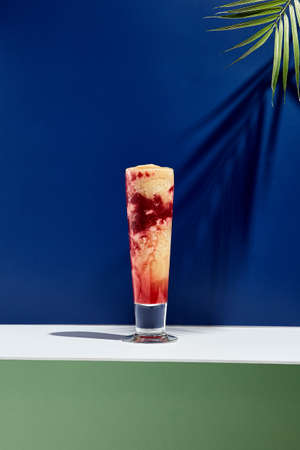 Raspberry and banana smoothie over white and blue background. Palm leaf with sunshine and hard shadow. Fruit yogurt or shake drink in high glass. Summer, healthy, fresh drink conceptの写真素材