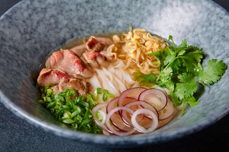 Vietnamese Cuisine - Beef Pho Noodle Soup or Pho Bo Soup. Served with Fresh Greens in Blue Bowl on Textured Dark Gray Table. Pho Bo Soup with Spicy Saucesの写真素材