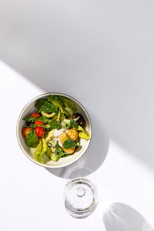 Salad bowl with avocado, tomato and cheese balls. Healthy vegetarian lunch with water glass on white...