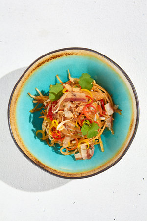 Stir fry noodles with chicken and bacon in Asian style. Fried udon with meat and vegetables in ceramic bowl on white background and sunlights shadows. Asian food in modern styleの写真素材