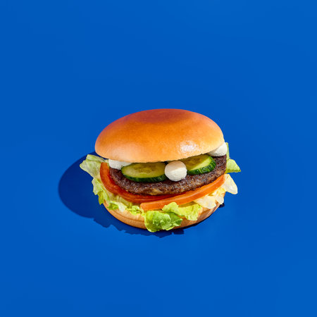 Tantalizing burger with feta cheese, fresh cucumbers, tomatoes, white sauce, and a meat patty on a blue background. Contemporary minimalist food photography, great for takeout. Square, low-angleの写真素材