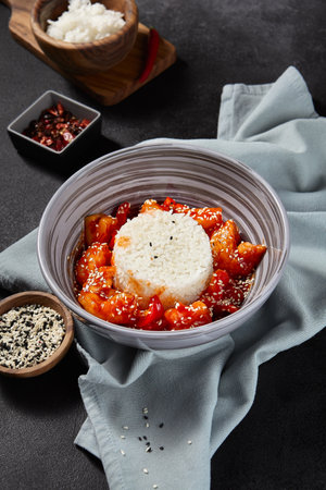 Aesthetic composition of fish and rice in a sweet and sour sauce with sesame in a gray bowl on a black textured background. Sesame and red pepper in a wooden bowl surround the dish. side viewの写真素材