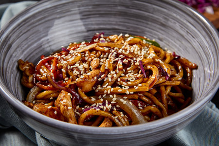 Side view, horizontal shot of a delicious udon dish stir-fried with chicken and vegetables in a savory garlic sauce. Chopsticks and a textured cloth complement the black background setting.の写真素材