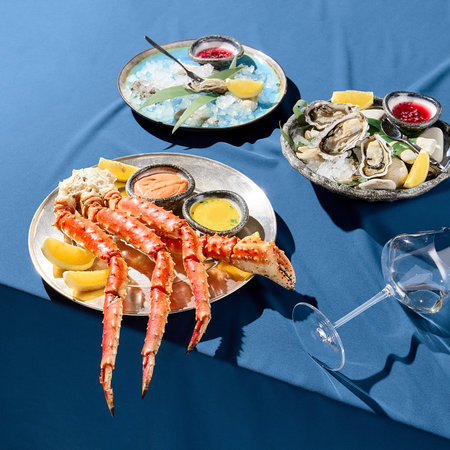 A square frame of a luxurious seafood platter with crab phalanges, lemon slices, sauces, and fresh oysters on ice. Served on a blue tablecloth against a uniform blue backdrop with stark shadows.の写真素材
