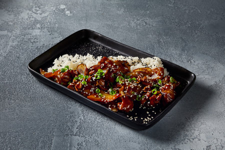 Horizontal side-view of the Kung Pao chicken, emphasizing the vibrant colors and texture on the black plate. The sweet-sour sauce glistens against the serene gray backdrop.の写真素材