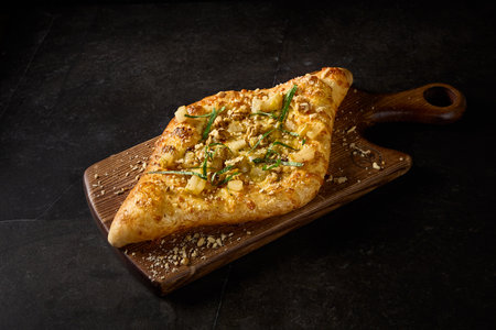 Side perspective of an innovative Adjarian khachapuri adorned with pear, nuts, and cheese, served on a wooden platter against a contrasting black setting.の写真素材