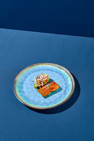 Diagonal side view vertical shot of cod in unagi sauce accompanied by grilled avocado and vegetables, elegantly presented on a blue ceramic plate, minimalist style with shadows on a blue backdrop.の写真素材