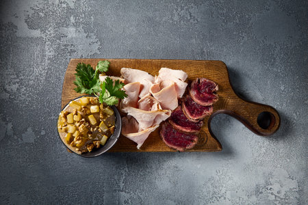Overhead view of a meat platter featuring roast beef, turkey pastrami, and chicken pÃ¢tÃ©, elegantly presented on a wooden board against a gray concrete backdrop.の写真素材