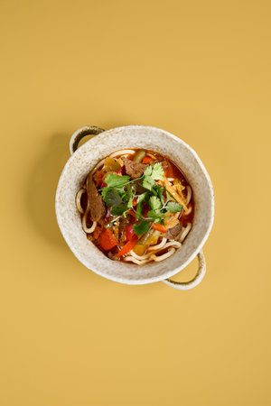 An appetizing top-down image of Lagman with noodles, beef, bell peppers, and garnish, served in a textured bowl on a yellow backdrop.の写真素材