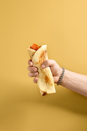 A person's hand displaying a freshly rolled Turkish Lahmacun pizza wrap with a ripe tomato peeking out, against a warm yellow background.の写真素材