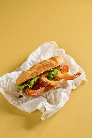A vertical view of a shrimp sandwich with crisp lettuce and tomato in a bread roll, wrapped in parchment on a sunny yellow surface.の写真素材