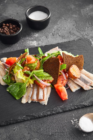 Lightly salted herring and potato salad garnished with fresh vegetables, served on a slate board against a textured black backgroundの写真素材