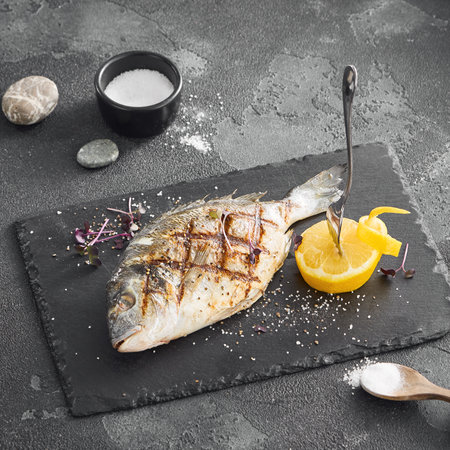 Perfectly grilled whole fish served with a slice of lemon on a slate board, highlighted against a textured gray backdrop, square composition.の写真素材