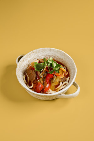 This vertical image showcases a bowl of traditional Lagman soup with a focus on the combination of noodles, sliced beef, bell peppers, and other vegetables, topped with fresh cilantro and sesame seedsの写真素材