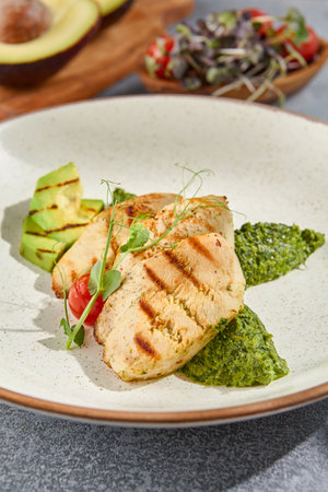Macro vertical side view capturing the textures of grilled chicken fillet, avocado, and spinach on a ceramic plate. Set against a gray concrete background with hard shadows.の写真素材