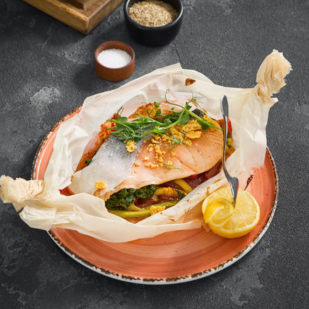 Freshly baked salmon steak on a colorful bed of vegetables, served in parchment paper on a terracotta plate with lemon wedges.の写真素材