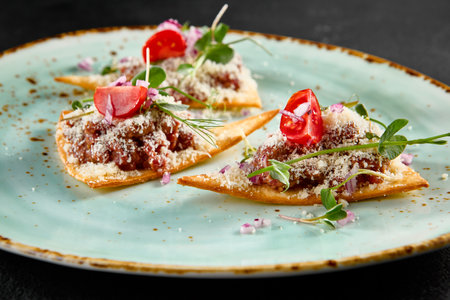 Elegant beef tartare on crispy toasts with parmesan, greens, and tomatoes on a speckled turquoise plate.の写真素材