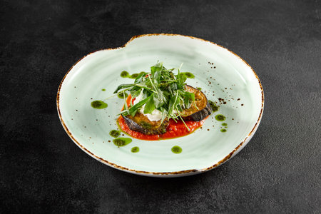 Baked eggplant with tomatoes and cheese, garnished with fresh greens on a ceramic plate.の写真素材