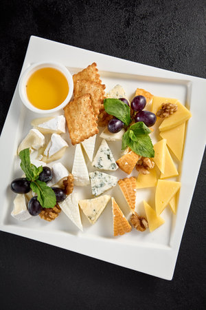 Top view of a cheese board with grapes and honey on a white plate against a black background.の写真素材