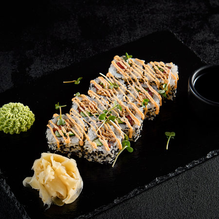 Sushi roll with vegetables and fried salmon inside, sesame seeds outside, and spicy sauce, presented on a black slate with soy sauce, wasabi, and ginger.の写真素材