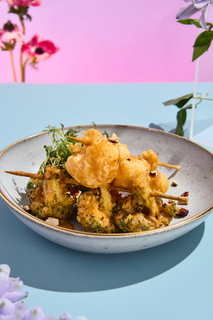 Elegant tempura vegetables on skewers with nut sauce amidst a surreal pastel floral backdrop.の写真素材
