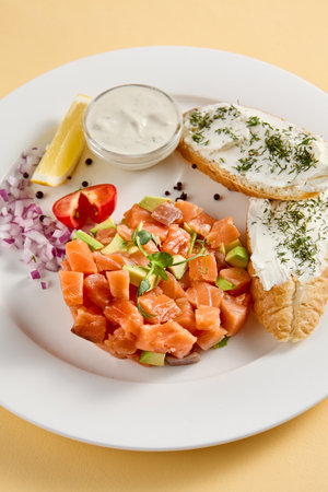 Salmon and avocado tartare with crispy bread and cream cheese, elegantly plated with lemon and dill on a white plate against a yellow background.の写真素材