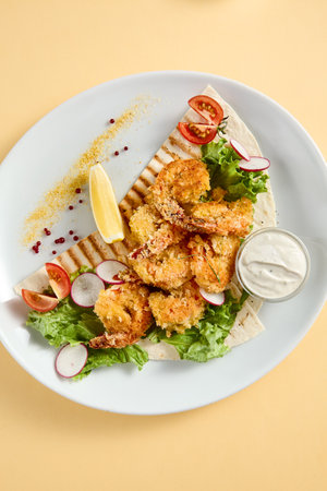 Tempura shrimp with tartar sauce on a bed of lettuce, presented on a white plate with lemon and tomato accents, against a soft yellow backdrop.の写真素材