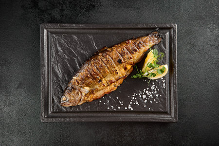 Grilled rainbow trout with a lemon wedge and fresh herbs on a dark stone plate.の写真素材
