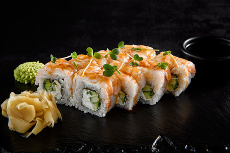 Shrimp roll with shrimp on top and cucumber and cheese inside, on a black slate with soy sauce, wasabi, and ginger.の写真素材