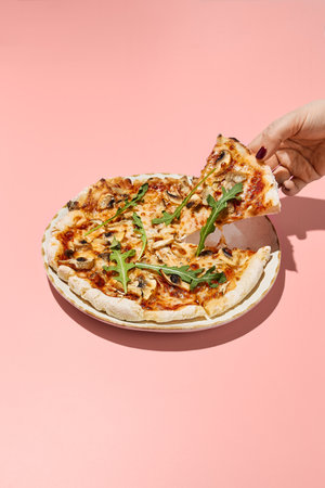 Woman's hand serving a slice of gluten-free pizza with mushrooms and tomatoes on a pink background.の写真素材
