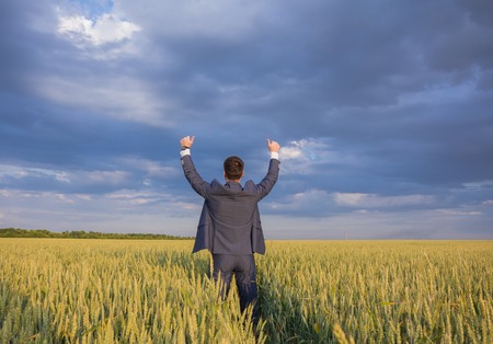 happy farmer, businessman, standing in wheat field over wind turbines background with his hands up and thumbs upの写真素材