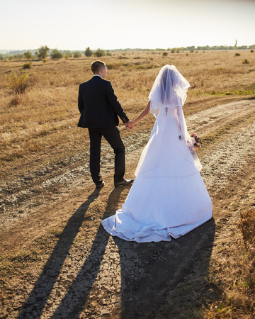 The bride and groom go through the field hand in hand. Happy bride and groom holding hands and walking in field on wedding day.の写真素材