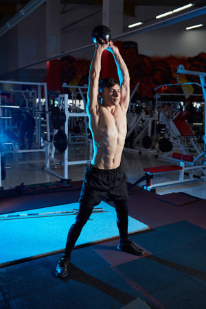 Young muscular man with naked torso working out in gym. Athletic male adult exercising with kettle bell. Fitness, sports concept. Blue backlightの写真素材