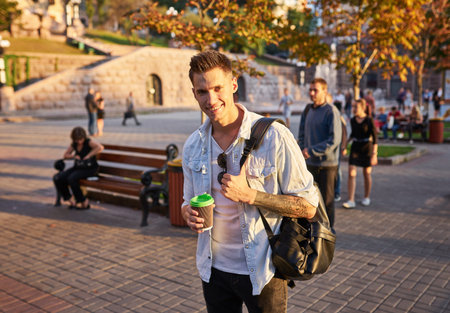 Hipster man standing with takeaway coffee, smiling plesantly, walking on th city street. Happy...