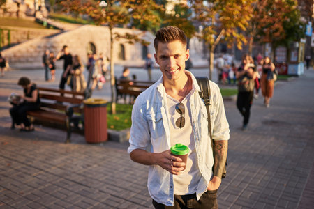 Hipster man standing with takeaway coffee, smiling plesantly, walking on th city street. Happy carefree handsome guy in eyewear. Millennial generationの写真素材