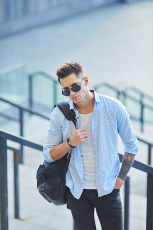 A young man in sunglasses with a black backpack stands on the stairs leaning on the railing.の写真素材