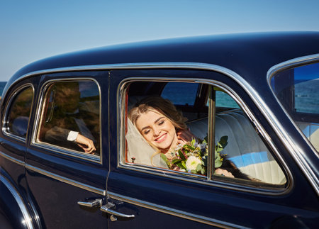 Pretty bride in luxury wedding dress sitting inside of retro car and holding wedding bouquetの写真素材
