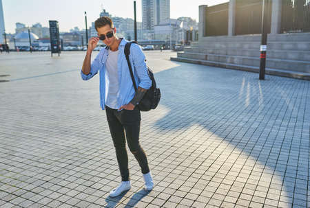 Fashionable young man in sunglasses with a black backpack walking along a city streetの写真素材